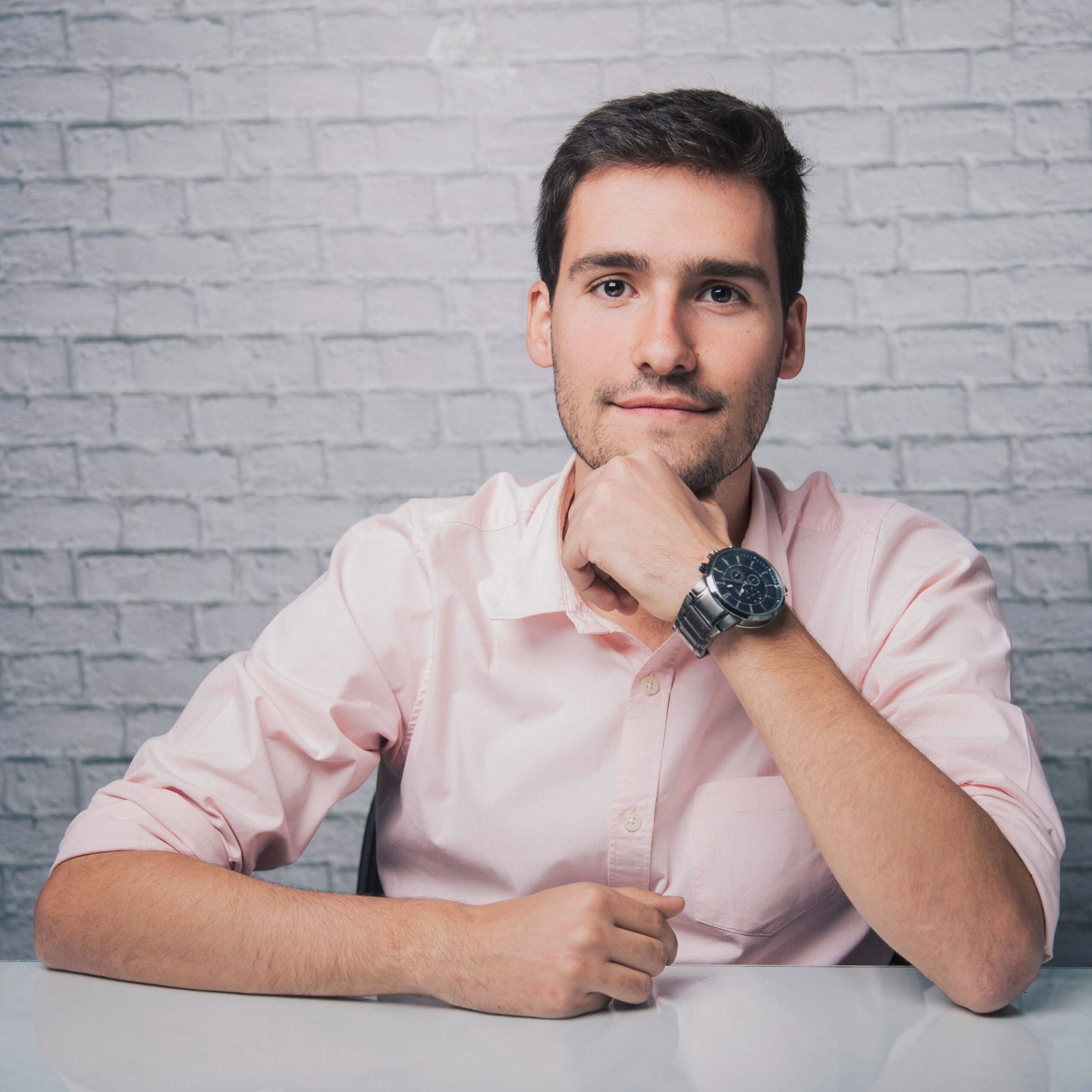 Positive young male in shirt with wristwatch sitting at desk with hand at chin while smiling and looking at camera in room with white brick wall